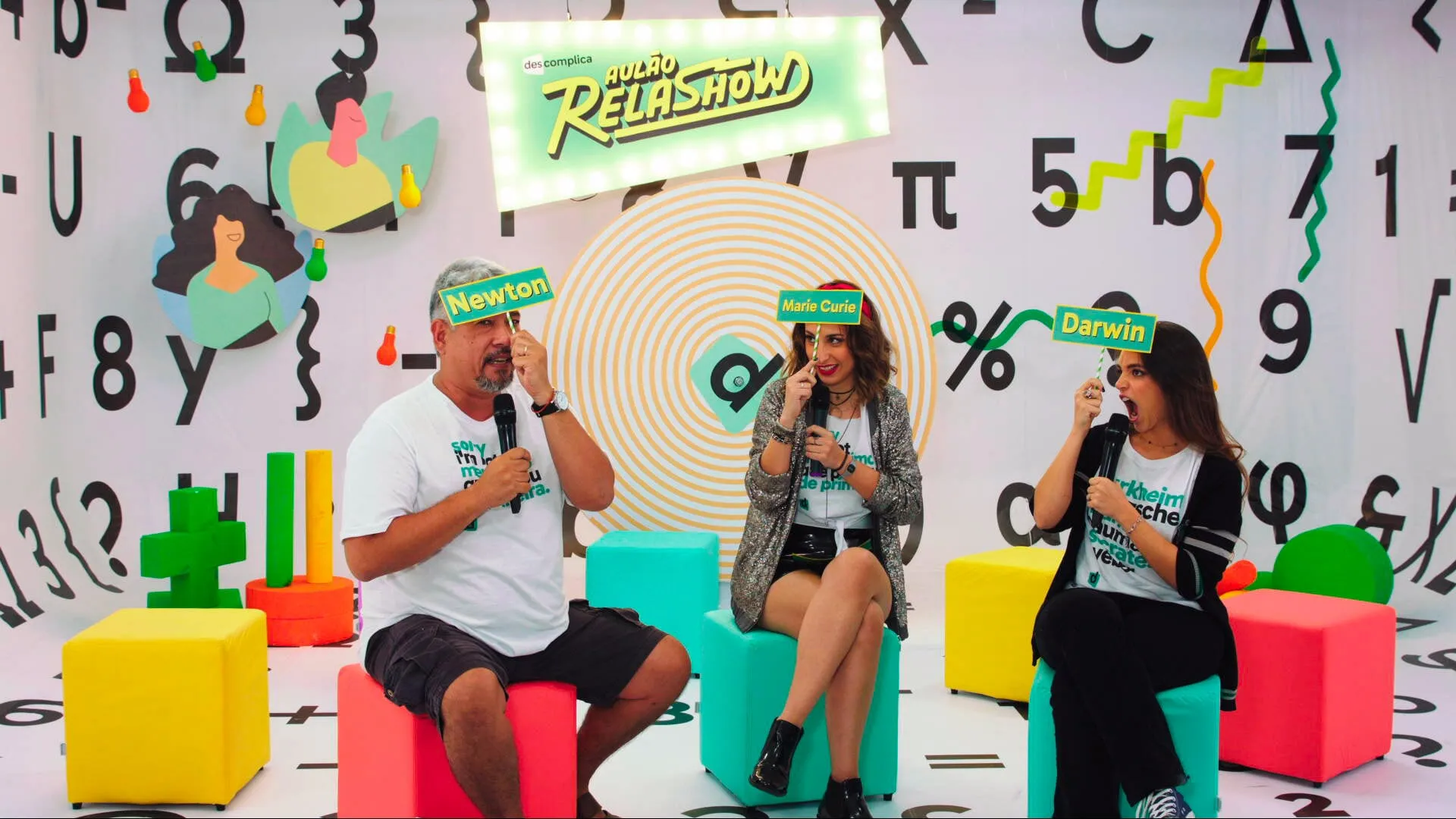 Descomplica's playful RelaShow study event, three hosts on a maths-symbol set holding scientist name cards